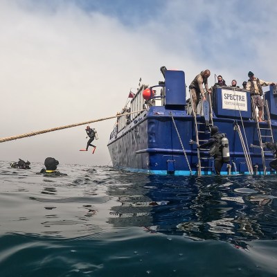Divers enter and exit a blue boat in the ocean, with one mid-air jumping from a platform.