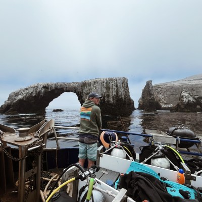 Person on boat with diving gear, overlooking rocky arch formation in calm sea.