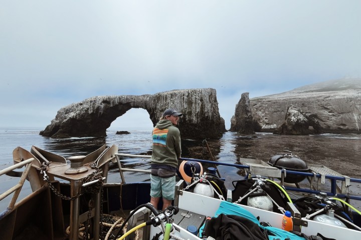 Person on boat with diving gear, overlooking rocky arch formation in calm sea.