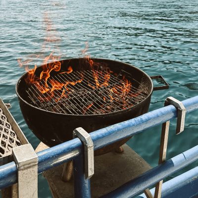 Burning grill on a boat with rocky island and lighthouse in the background.