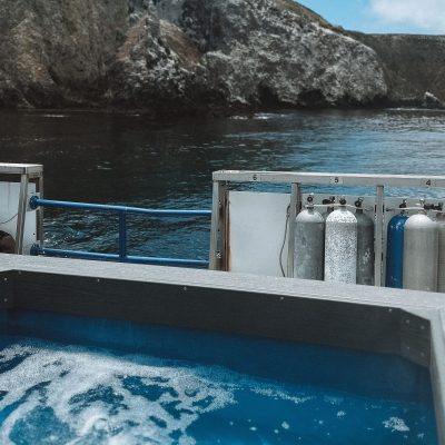 A hot tub on a boat with scuba tanks by rocky coastal cliffs under a cloudy sky.