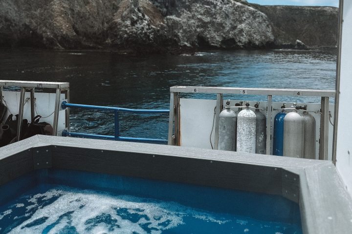 A hot tub on a boat with scuba tanks by rocky coastal cliffs under a cloudy sky.