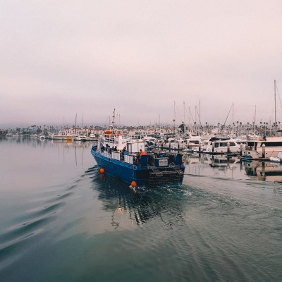 Blue boat sailing through a marina with many docked yachts under a cloudy sky.