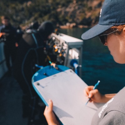 Person on boat with clipboard, writing, while others prepare diving equipment.