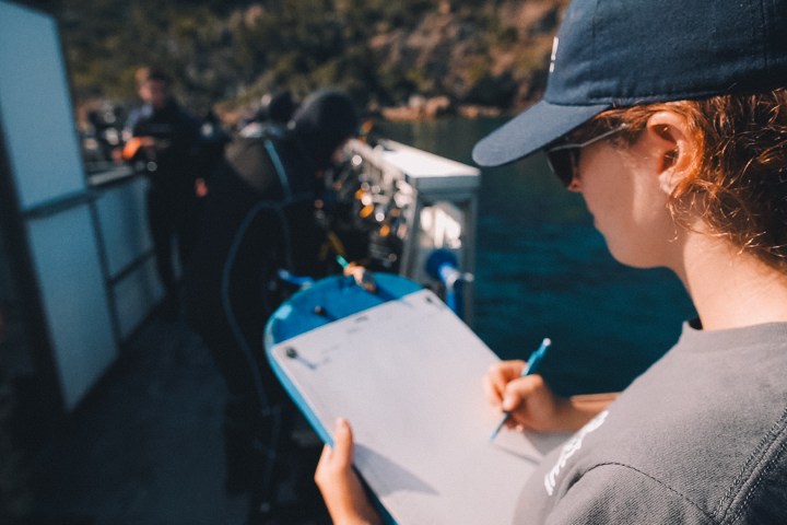 Person on boat with clipboard, writing, while others prepare diving equipment.