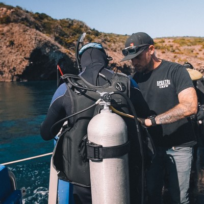 Diver in wetsuit with scuba gear on a boat, preparing to dive near rocky coastline.