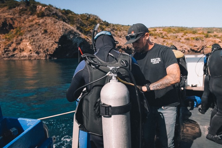 Diver in wetsuit with scuba gear on a boat, preparing to dive near rocky coastline.