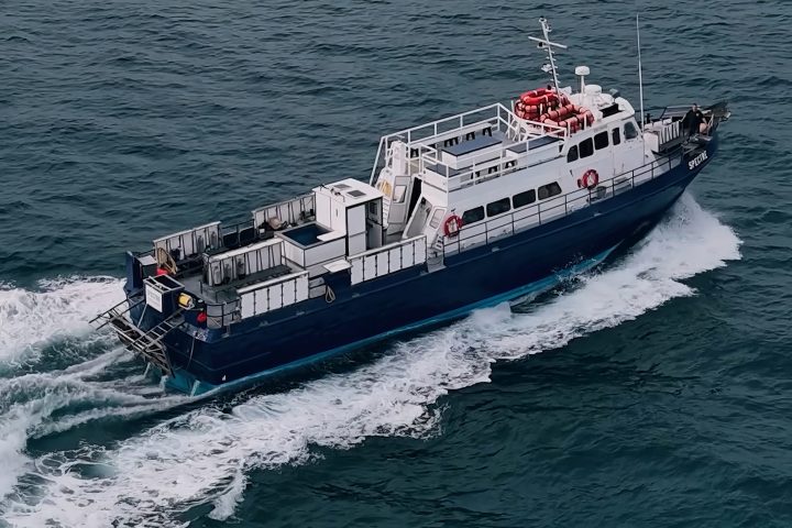 Blue and white boat sailing on a calm sea with a mountainous coastline in the distance.